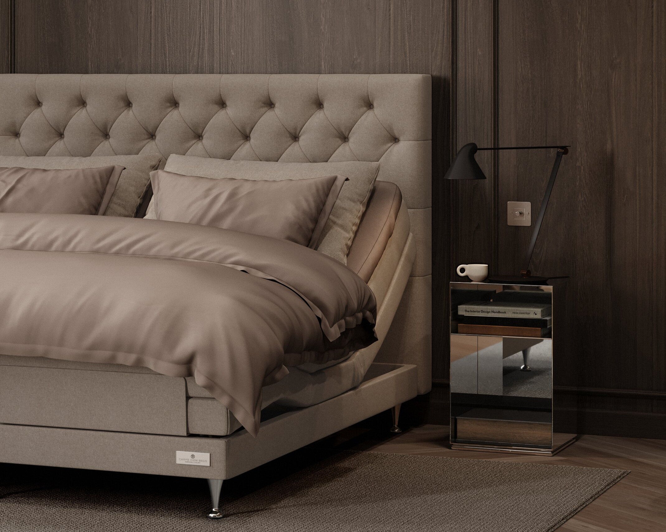 A modern bedroom scene featuring a bed with a tufted beige upholstered headboard, matching beige bedding, a dark wood-paneled wall, a small mirrored nightstand with a black lamp, and a gray rug.