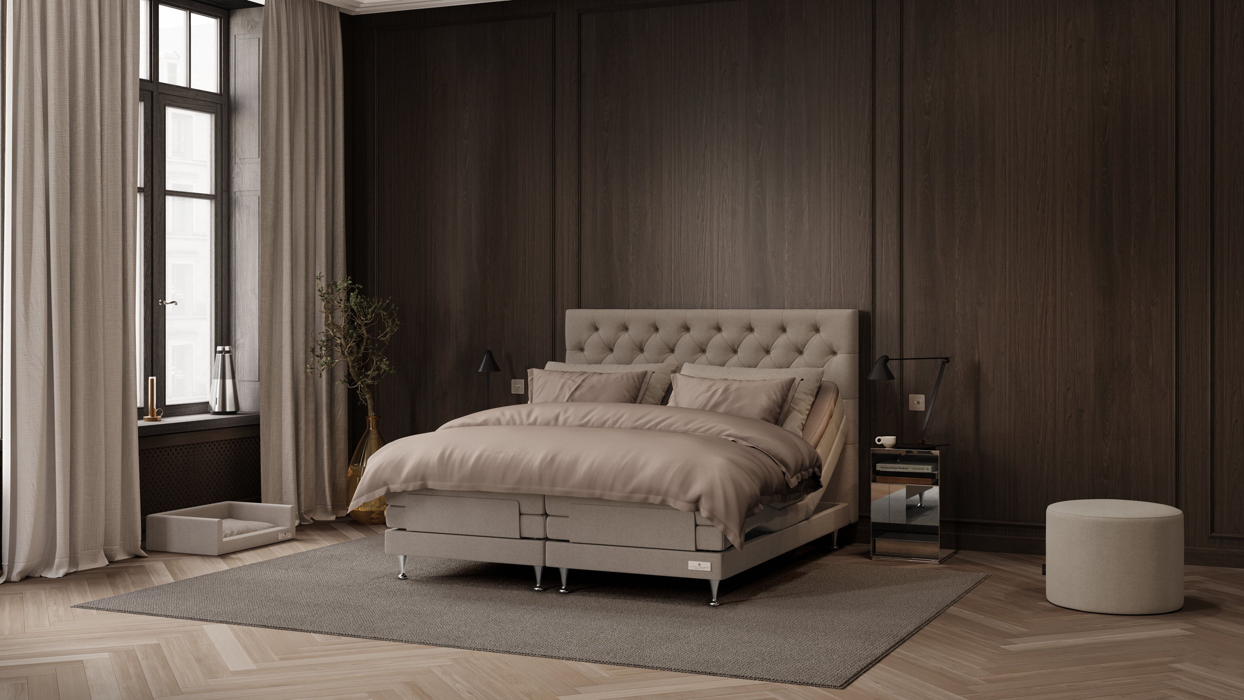 A modern bedroom featuring a tufted beige platform bed with matching bedding, set against a dark wood-paneled wall with a window, curtains, side tables, and a round ottoman.