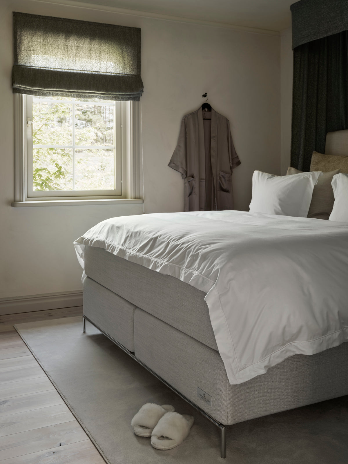A modern bedroom featuring a low-profile platform bed with a light gray upholstered frame and crisp white bedding. A matching gray robe hangs on a wall hook beside a window with a gray roman blind, and a pair of white slippers sits on the floor nearby.